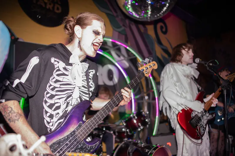 A bassist in a skeleton costume and white face paint sings mid-song beside a guitarist dressed as a ghost, under a disco ball at Junkyard Social Club